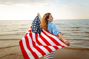 Amerikan bayrağı taşıyan mutlu kadın deniz kıyısında eğleniyor. Genç turist kendini özgür hissediyor ve Amerika 'nın Bağımsızlık Günü' nü kutluyor. 4 Temmuz kavramı, rahatlama, keyif.
