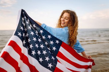 Amerikan bayrağı taşıyan mutlu kadın deniz kıyısında eğleniyor. Genç turist kendini özgür hissediyor ve Amerika 'nın Bağımsızlık Günü' nü kutluyor. 4 Temmuz kavramı, rahatlama, keyif.