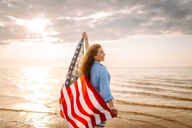 Amerikan bayrağı taşıyan mutlu kadın deniz kıyısında eğleniyor. Genç turist kendini özgür hissediyor ve Amerika 'nın Bağımsızlık Günü' nü kutluyor. 4 Temmuz kavramı, rahatlama, keyif.