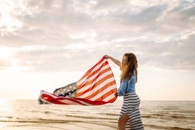 Amerikan bayrağı taşıyan mutlu kadın deniz kıyısında eğleniyor. Genç turist kendini özgür hissediyor ve Amerika 'nın Bağımsızlık Günü' nü kutluyor. 4 Temmuz kavramı, rahatlama, keyif.