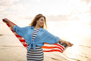 Amerikan bayrağı taşıyan mutlu kadın deniz kıyısında eğleniyor. Genç turist kendini özgür hissediyor ve Amerika 'nın Bağımsızlık Günü' nü kutluyor. 4 Temmuz kavramı, rahatlama, keyif.
