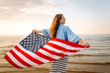 Amerikan bayrağı taşıyan mutlu kadın deniz kıyısında eğleniyor. Genç turist kendini özgür hissediyor ve Amerika 'nın Bağımsızlık Günü' nü kutluyor. 4 Temmuz kavramı, rahatlama, keyif.