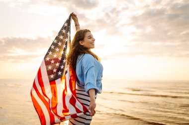 Amerikan bayrağı taşıyan mutlu kadın deniz kıyısında eğleniyor. Genç turist kendini özgür hissediyor ve Amerika 'nın Bağımsızlık Günü' nü kutluyor. 4 Temmuz kavramı, rahatlama, keyif.