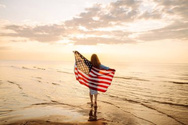 Amerikan bayrağı taşıyan mutlu kadın deniz kıyısında eğleniyor. Genç turist kendini özgür hissediyor ve Amerika 'nın Bağımsızlık Günü' nü kutluyor. 4 Temmuz kavramı, rahatlama, keyif.