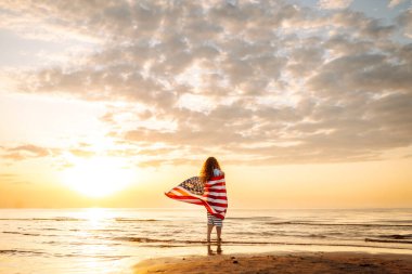 Amerikan bayrağı taşıyan mutlu kadın deniz kıyısında eğleniyor. Genç turist kendini özgür hissediyor ve Amerika 'nın Bağımsızlık Günü' nü kutluyor. 4 Temmuz kavramı, rahatlama, keyif.