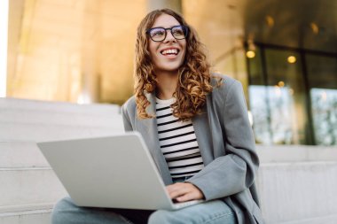 Günbatımı iş merkezine karşı dizüstü bilgisayarı olan ceketli neşeli bir kadın. Dışarıda dizüstü bilgisayarda çalışan bir iş kadını. Serbest konsept, boş zaman. Yaşam biçimi.