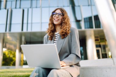 Günbatımı iş merkezine karşı dizüstü bilgisayarı olan ceketli neşeli bir kadın. Dışarıda dizüstü bilgisayarda çalışan bir iş kadını. Serbest konsept, boş zaman. Yaşam biçimi.