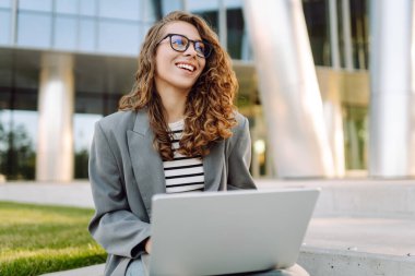 Günbatımı iş merkezine karşı dizüstü bilgisayarı olan ceketli neşeli bir kadın. Dışarıda dizüstü bilgisayarda çalışan bir iş kadını. Serbest konsept, boş zaman. Yaşam biçimi.