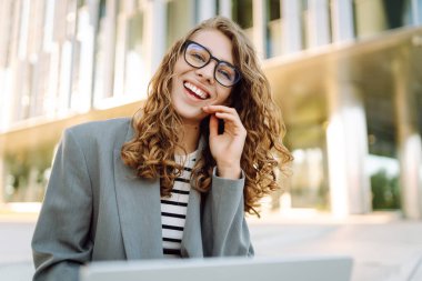 Günbatımı iş merkezine karşı dizüstü bilgisayarı olan ceketli neşeli bir kadın. Dışarıda dizüstü bilgisayarda çalışan bir iş kadını. Serbest konsept, boş zaman. Yaşam biçimi.