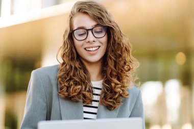 Günbatımı iş merkezine karşı dizüstü bilgisayarı olan ceketli neşeli bir kadın. Dışarıda dizüstü bilgisayarda çalışan bir iş kadını. Serbest konsept, boş zaman. Yaşam biçimi.