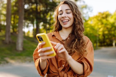 Elinde telefonla parkta yürüyen genç bir kadının portresi. Telefona bakan, dışarıda mesajlaşan güzel bir kadın. Bloglama, yürüme konsepti.