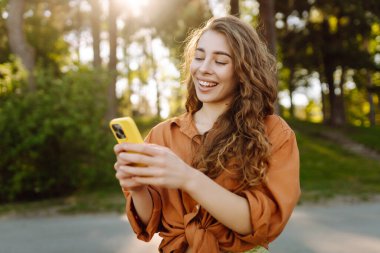 Elinde telefonla parkta yürüyen genç bir kadının portresi. Telefona bakan, dışarıda mesajlaşan güzel bir kadın. Bloglama, yürüme konsepti.