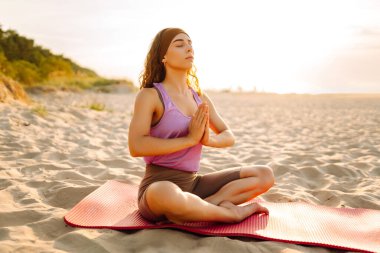 Arka planda deniz olan parlak minder üzerinde yoga yapan bir kadın. Zene yakalayan ve gün batımında plajda dinlenen sportif bir kadın. Rahatlama kavramı, keyif. Etkin yaşam biçimi.