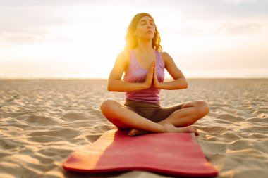 Arka planda deniz olan parlak minder üzerinde yoga yapan bir kadın. Zene yakalayan ve gün batımında plajda dinlenen sportif bir kadın. Rahatlama kavramı, keyif. Etkin yaşam biçimi.