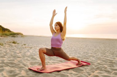 Arka planda deniz olan parlak minder üzerinde yoga yapan bir kadın. Zene yakalayan ve gün batımında plajda dinlenen sportif bir kadın. Rahatlama kavramı, keyif. Etkin yaşam biçimi.