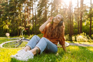 Kulaklıklı mutlu kadın yeşil bir çayırda oturuyor kulaklıkları başında, arka planda bir bisiklet yürüyor. Kadın güneşli bir parkta bisiklet sürdükten sonra dinleniyor. Etkin yaşam biçimi. 