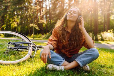 Kulaklıklı mutlu kadın yeşil bir çayırda oturuyor kulaklıkları başında, arka planda bir bisiklet yürüyor. Kadın güneşli bir parkta bisiklet sürdükten sonra dinleniyor. Etkin yaşam biçimi. 