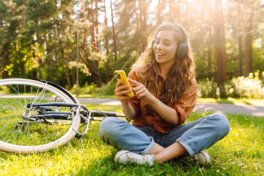 Kulaklıklı mutlu kadın yeşil bir çayırda oturuyor kulaklıkları başında, arka planda bir bisiklet yürüyor. Kadın güneşli bir parkta bisiklet sürdükten sonra dinleniyor. Etkin yaşam biçimi. 