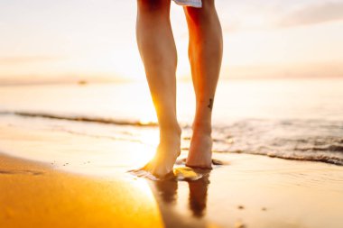 Gün batımı sahili boyunca deniz boyunca yürüyen bir kadının bacaklarının yakın çekimi. Elbise giymiş genç bir kadın açık havada güneşli havanın tadını çıkarır. Yürüme kavramı, keyif..