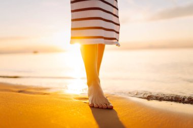 Gün batımı sahili boyunca deniz boyunca yürüyen bir kadının bacaklarının yakın çekimi. Elbise giymiş genç bir kadın açık havada güneşli havanın tadını çıkarır. Yürüme kavramı, keyif..