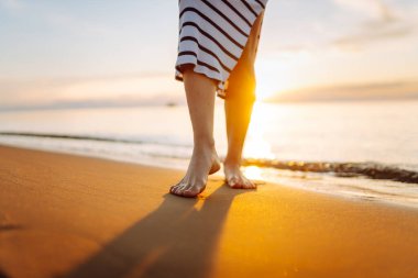 Gün batımı sahili boyunca deniz boyunca yürüyen bir kadının bacaklarının yakın çekimi. Elbise giymiş genç bir kadın açık havada güneşli havanın tadını çıkarır. Yürüme kavramı, keyif..