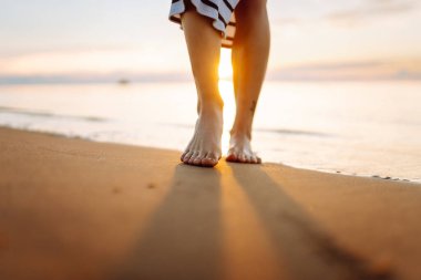 Gün batımı sahili boyunca deniz boyunca yürüyen bir kadının bacaklarının yakın çekimi. Elbise giymiş genç bir kadın açık havada güneşli havanın tadını çıkarır. Yürüme kavramı, keyif..