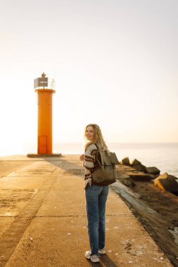 Arka planda deniz feneri olan mutlu bir kadın rıhtımda eğleniyor. Gün batımında özgür hisseden güzel bir kadın. Macera, yürüyüş konsepti. Yaşam biçimi.