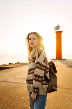 Arka planda deniz feneri olan mutlu bir kadın rıhtımda eğleniyor. Gün batımında özgür hisseden güzel bir kadın. Macera, yürüyüş konsepti. Yaşam biçimi.