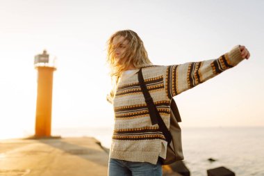 Arka planda deniz feneri olan mutlu bir kadın rıhtımda eğleniyor. Gün batımında özgür hisseden güzel bir kadın. Macera, yürüyüş konsepti. Yaşam biçimi.