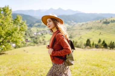 Sırt çantalı genç bir kadın yeşil bir tepede yürüyor. Şapkalı bir bayan gezgin güneşli havanın tadını çıkarıyor, özgür hissediyor. Macera konsepti, özgürlük. Etkin yaşam biçimi.