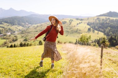 Sırt çantalı genç bir kadın yeşil bir tepede yürüyor. Şapkalı bir bayan gezgin güneşli havanın tadını çıkarıyor, özgür hissediyor. Macera konsepti, özgürlük. Etkin yaşam biçimi.