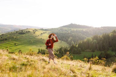 Sırt çantalı genç bir kadın yeşil bir tepede yürüyor. Şapkalı bir bayan gezgin güneşli havanın tadını çıkarıyor, özgür hissediyor. Macera konsepti, özgürlük. Etkin yaşam biçimi.