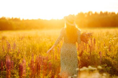 Gülümseyen kadın gün batımında mor çiçek tarlasında eğleniyor. Vahşi çiçek buketi olan güzel bir kadın doğanın tadını çıkarıyor. Rahatlama, poz verme konsepti.