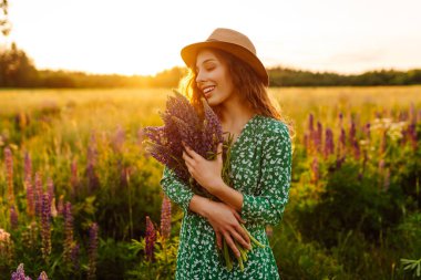 Gülümseyen kadın gün batımında mor çiçek tarlasında eğleniyor. Vahşi çiçek buketi olan güzel bir kadın doğanın tadını çıkarıyor. Rahatlama, poz verme konsepti.