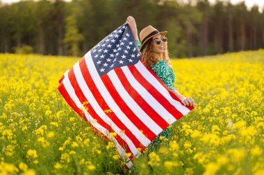 Parlak elbiseli genç bir kadın elinde Amerikan bayrağıyla çiçek tarlasında yürüyor. Gün batımında kolza tarlasında bayrağıyla özgürlük hisseden bir kadın. Bağımsızlık, yürüyüşler, eğlence konsepti.
