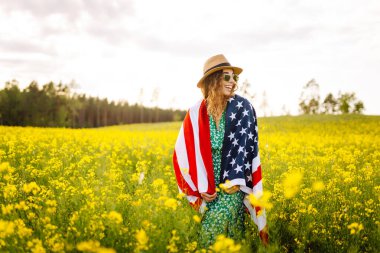 Parlak elbiseli genç bir kadın elinde Amerikan bayrağıyla çiçek tarlasında yürüyor. Gün batımında kolza tarlasında bayrağıyla özgürlük hisseden bir kadın. Bağımsızlık, yürüyüşler, eğlence konsepti.