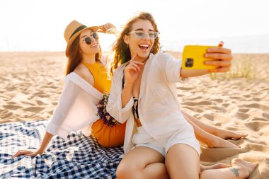 İki genç bayan arkadaş sahilde selfie çekiyorlar. Güzel kadınlar gün batımında buket ve telefonla eğleniyorlar. Eğlence, rahatlama, blog konsepti.