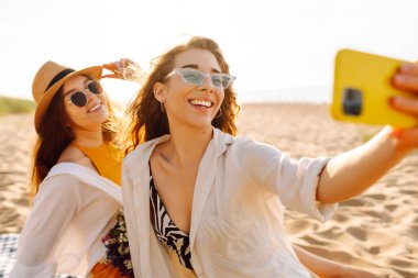 İki genç bayan arkadaş sahilde selfie çekiyorlar. Güzel kadınlar gün batımında buket ve telefonla eğleniyorlar. Eğlence, rahatlama, blog konsepti.