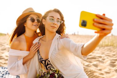 İki genç bayan arkadaş sahilde selfie çekiyorlar. Güzel kadınlar gün batımında buket ve telefonla eğleniyorlar. Eğlence, rahatlama, blog konsepti.