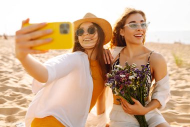 İki genç bayan arkadaş sahilde selfie çekiyorlar. Güzel kadınlar gün batımında buket ve telefonla eğleniyorlar. Eğlence, rahatlama, blog konsepti.