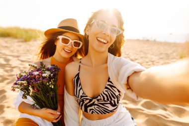 İki genç bayan arkadaş sahilde selfie çekiyorlar. Güzel kadınlar gün batımında buket ve telefonla eğleniyorlar. Eğlence, rahatlama, blog konsepti.