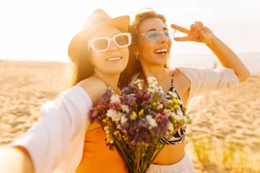 İki genç bayan arkadaş sahilde selfie çekiyorlar. Güzel kadınlar gün batımında buket ve telefonla eğleniyorlar. Eğlence, rahatlama, blog konsepti.