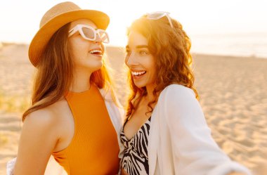 İki genç bayan arkadaş sahilde selfie çekiyorlar. Güzel kadınlar gün batımında buket ve telefonla eğleniyorlar. Eğlence, rahatlama, blog konsepti.