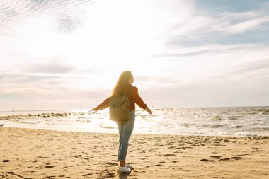 Sırt çantalı kadın gezginin gün batımında kumlu sahilde yürüyüşü. Deniz kıyısında özgürlük hisseden genç bir kadın. Özgürlük, seyahat kavramı.