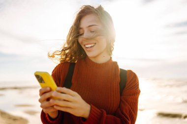 Fırtınalı bir denizin arka planında elinde telefonla gülümseyen bir kadın. Bir turist telefonuna mesaj atıyor ve saçlarında rüzgarın tadını çıkarıyor. Teknoloji, iletişim ve macera kavramı.