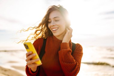 Fırtınalı bir denizin arka planında elinde telefonla gülümseyen bir kadın. Bir turist telefonuna mesaj atıyor ve saçlarında rüzgarın tadını çıkarıyor. Teknoloji, iletişim ve macera kavramı.