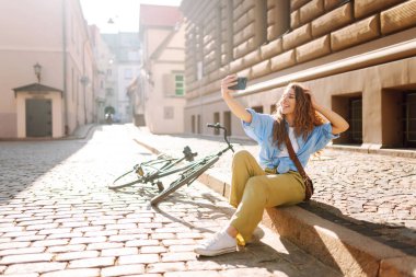 Bisikletli mutlu turist güneşli bir sokakta selfie çekiyor. Güzel bir kadın yürüyüş yapmaktan ve blog çekmekten hoşlanır. Turizm, eğlence konsepti.