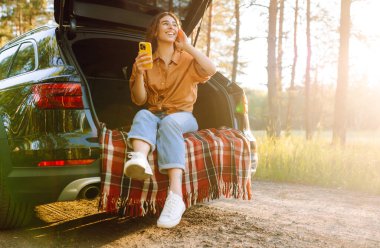 Mutlu kadın elinde telefonuyla bir arabanın bagajında oturuyor. Turistler açık havayı sever, özgürlüğü hisseder. Macera konsepti, tatil. Etkin yaşam biçimi.