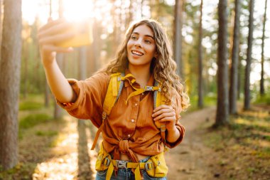 Güneşli bir ormanda selfie çeken sırt çantalı güzel bir kadın yürüyüşçünün portresi. Yürüyüş ve blog yapmaktan hoşlanan genç bir kadın. Macera konsepti. Etkin yaşam biçimi.
