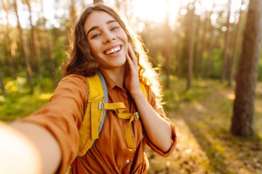 Güneşli bir ormanda selfie çeken sırt çantalı güzel bir kadın yürüyüşçünün portresi. Yürüyüş ve blog yapmaktan hoşlanan genç bir kadın. Macera konsepti. Etkin yaşam biçimi.